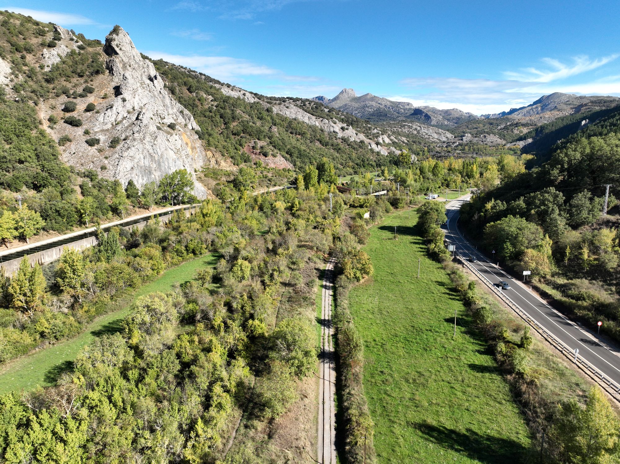 Dos raíles avanzan por una franja recta entre prados hacia el entorno del puente; a la izquierda roquedo y canal elevado, a la derecha carretera.