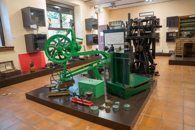 Vista general desde lado opuesto: máquina verde, báscula de plataforma, dos balanzas pequeñas, juegos de pesas y botiquín verde; máquina negra detrás.