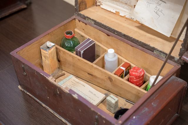 Detalle oblicuo del interior de botiquín de madera marrón con compartimentos, frascos, cajas, instrumental y documentos; tapa parcialmente fuera de marco.